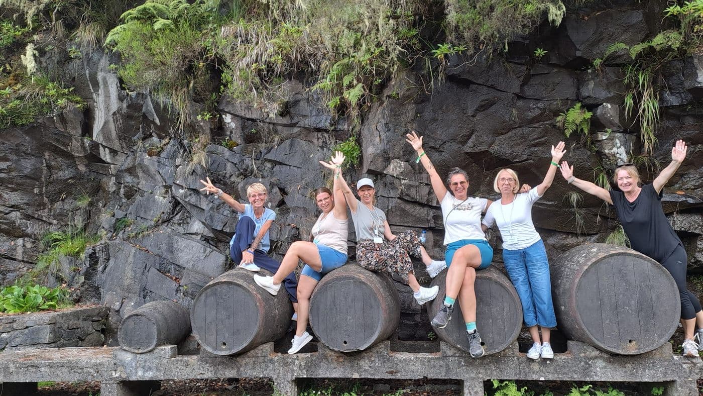 Sieben Vertreterinnen von HOLIDAY LAND Reisebüros sitzen und stehen lachend auf großen Holzfässern vor einer bewachsenen Felswand und strecken die Arme in die Höhe.