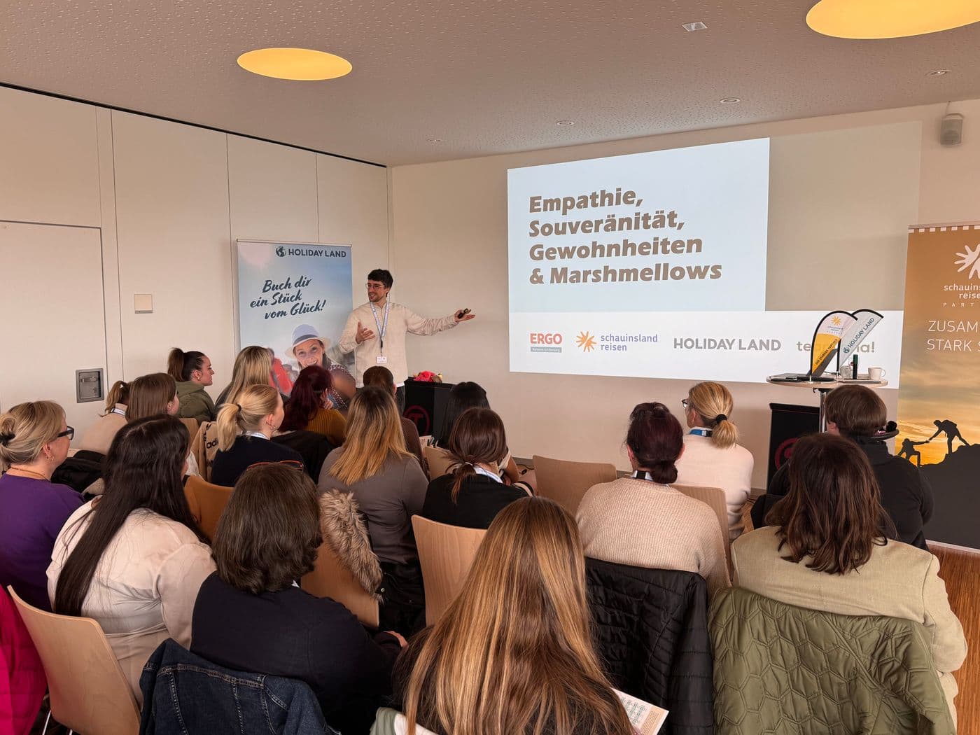 Eine größere Gruppe sitzt in einem Seminarraum und hört einem Vortragenden zu. Auf der Leinwand steht der Titel „Empathie, Souveränität, Gewohnheiten & Marshmellows“, darunter Logos von ERGO, schauinsland-reisen und HOLIDAY LAND. Der Vortragende steht vorn neben der Leinwand und gestikuliert, während hinter ihm ein Roll-Up mit dem Slogan „Buch dir ein Stück vom Glück!“ zu sehen ist. Die Teilnehmenden sitzen in Reihen und schauen nach vorn zur Präsentation.