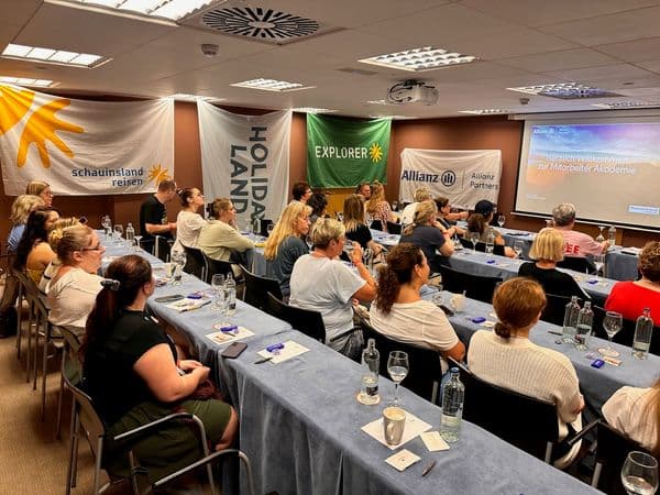Teilnehmende der HOLIDAY LAND Mitarbeiterakademie auf Gran Canaria verfolgen eine Schulung im Seminarraum. Banner von HOLIDAY LAND, schauinsland-reisen, EXPLORER und Allianz Partners sind im Hintergrund zu sehen.