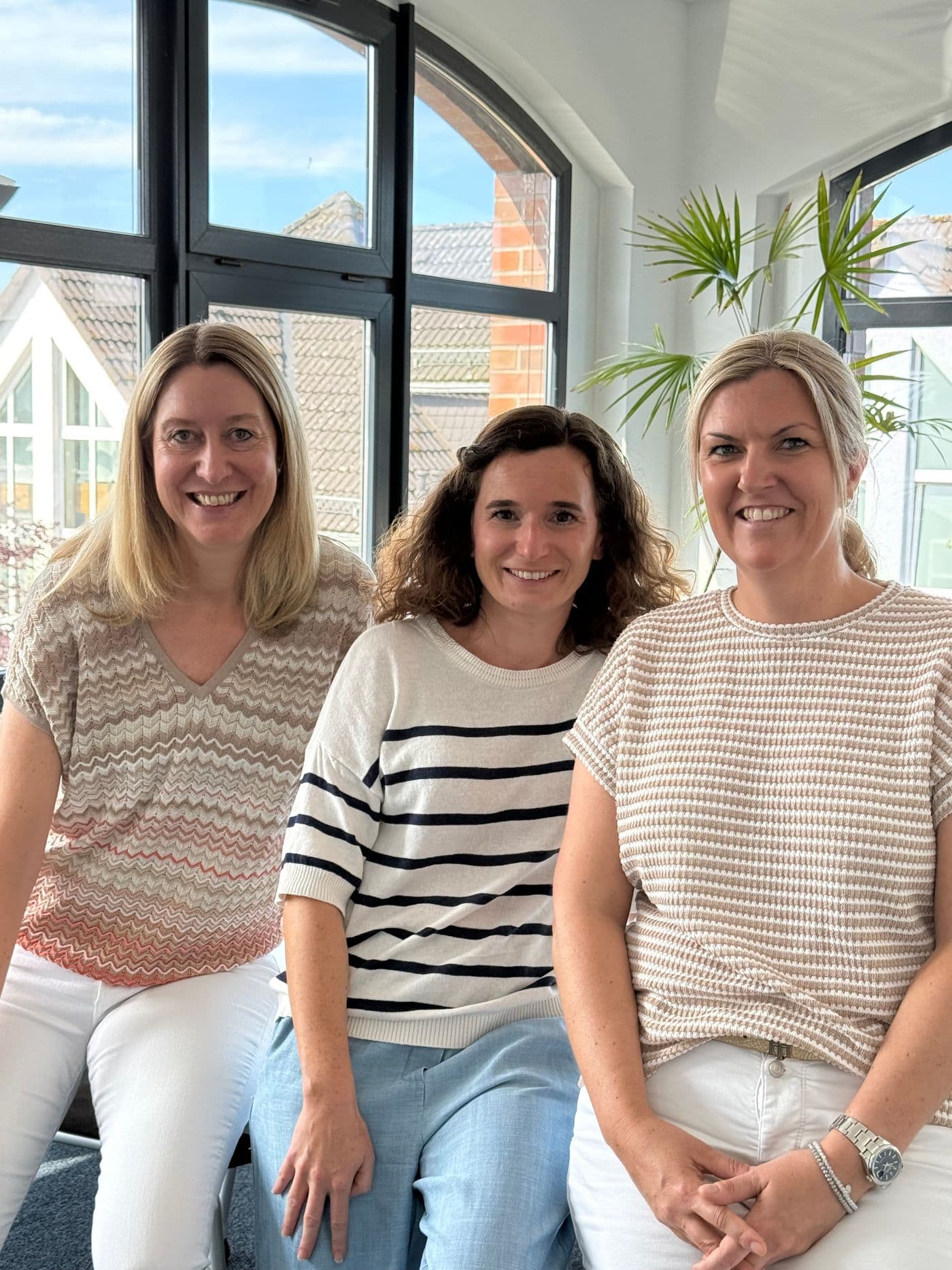 Silke Hülsewig, Helen Schröder und Nina Baumann sitzen an einem sonnigen Platz im Büro nebeneinander und lächeln in die Kamera.
